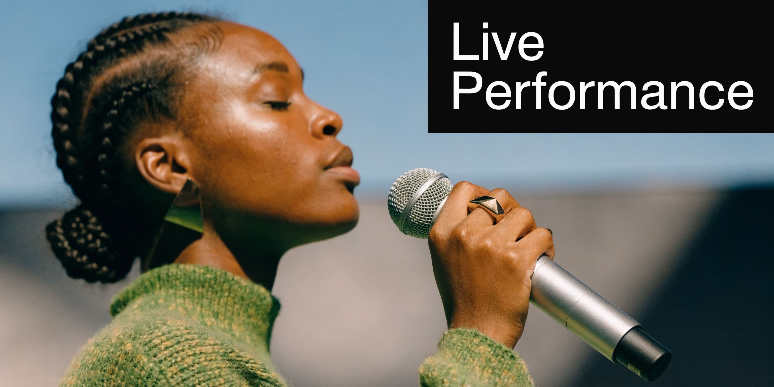 A Black woman with braided hair wearing a green sweater singing into a microphone outdoors.