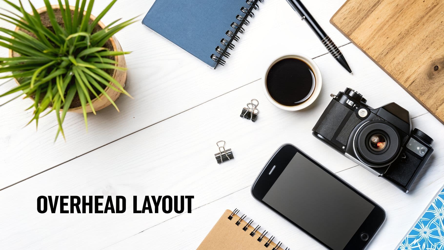 Overhead shot of a clean white wooden desk with a plant, coffee, camera, smartphone, and notebooks.