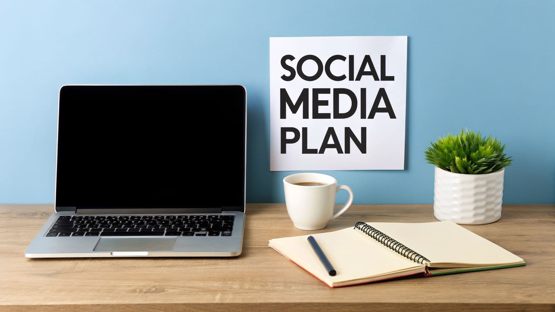 Workspace with a laptop, coffee, notebook, plant, and a 'SOCIAL MEDIA PLAN' sign.